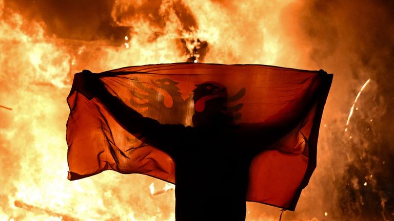 Ein Protestant hält am15.10.2026 während der Proteste gegen die Regierung in Tirana vor einem bennenden Kiosk die albanische Flagge hoch.