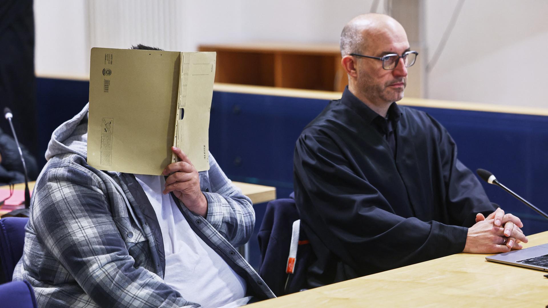 In einem Gerichtssaal sitzt der Hauptangeklagte links an einem Tisch. Er hält eine aufgeklappte Akte vor sein Gesicht. Rechts neben ihm sitzt sein Anwalt in schwarzer Robe.