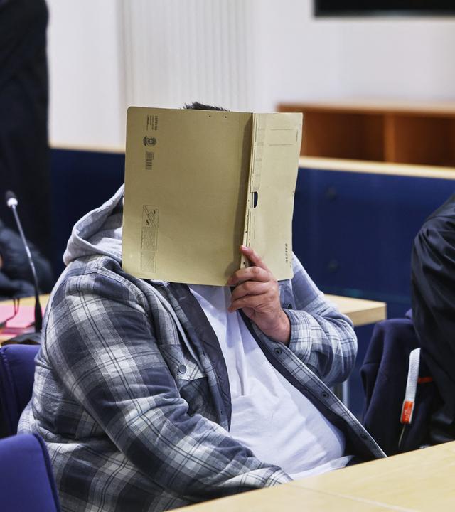 In einem Gerichtssaal sitzt der Hauptangeklagte links an einem Tisch. Er hält eine aufgeklappte Akte vor sein Gesicht. Rechts neben ihm sitzt sein Anwalt in schwarzer Robe.