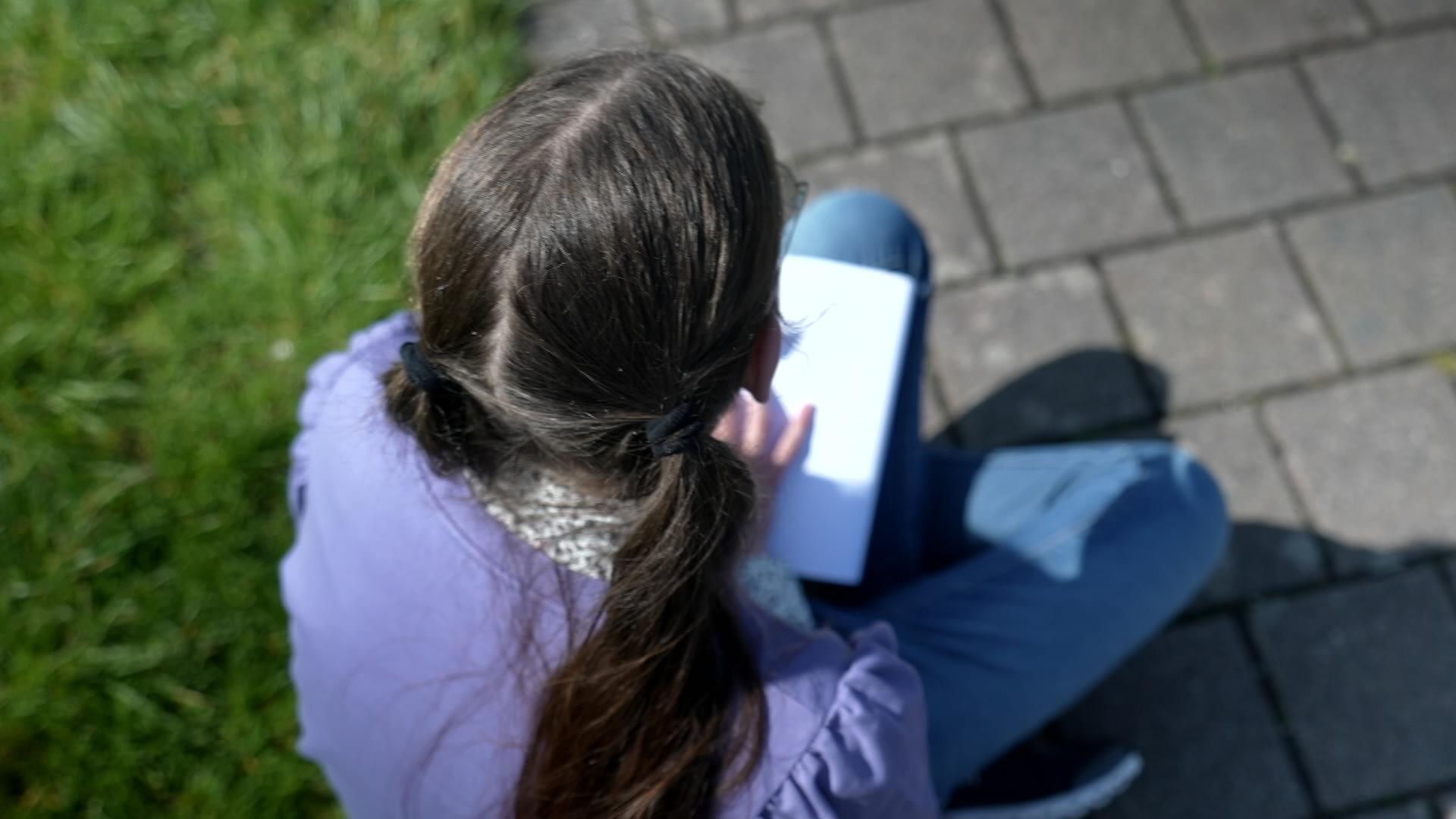 Mädchen mit Zöpfen (Rückansicht) sitzt auf dem Rasen und schreibt in Notizbuch