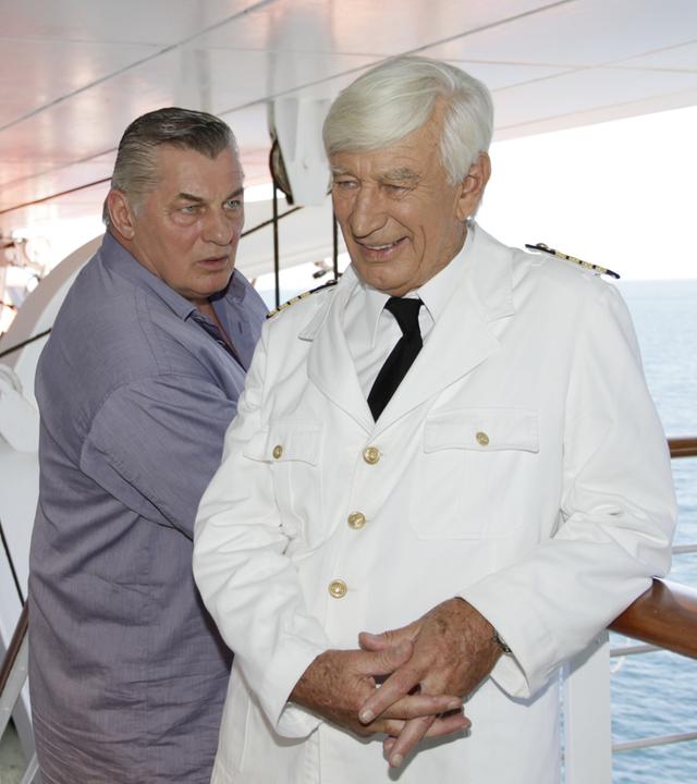 "Das Traumschiff: Puerto Rico": Paul (Heinz Hoenig) steht an der Reling der MS Amadea und blickt fragend Kapitän Paulsen (Siegfried Rauch) an. Dieser lächelt und hält die Hände gefaltet.
