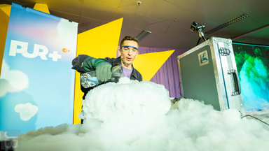 PUR+-Moderator Eric Mayer steht mit Schutzbrille und Handschuhen vor einer großen Wolke aus weißem Nebel. Er gießt eine klare Flüssigkeit aus einer Flasche in einen Behälter, der vom Nebel weitgehend verdeckt ist. Im Hintergrund sind das PUR+-Logo und gelbe Bühnenelemente zu sehen