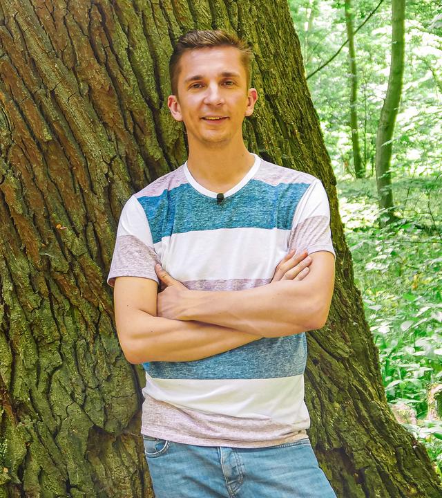 PUR+ Moderator Eric Mayer steht mit verschränkten Armen vor einem Baum. Zu sehen ist Eric vor einen sehr breiten Baumstamm.