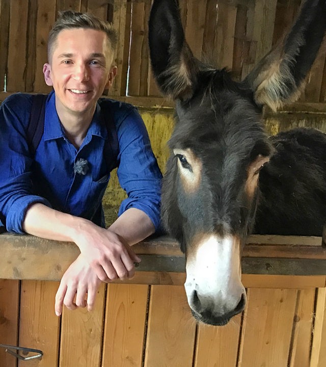 PUR+ Moderator Eric Mayer steht in einem Stall und lehnt an der Brüstung. Rechts und links von ihm steht je ein Esel.