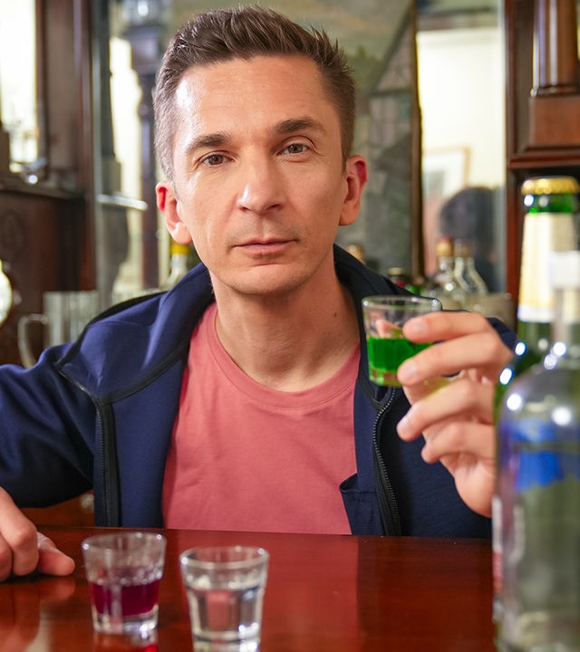 Eric sitzt an einer Bar und hält ein Glas mit Alkohol gefüllt in der Hand. Vor ihm stehen weitere Gläser und Flaschen.