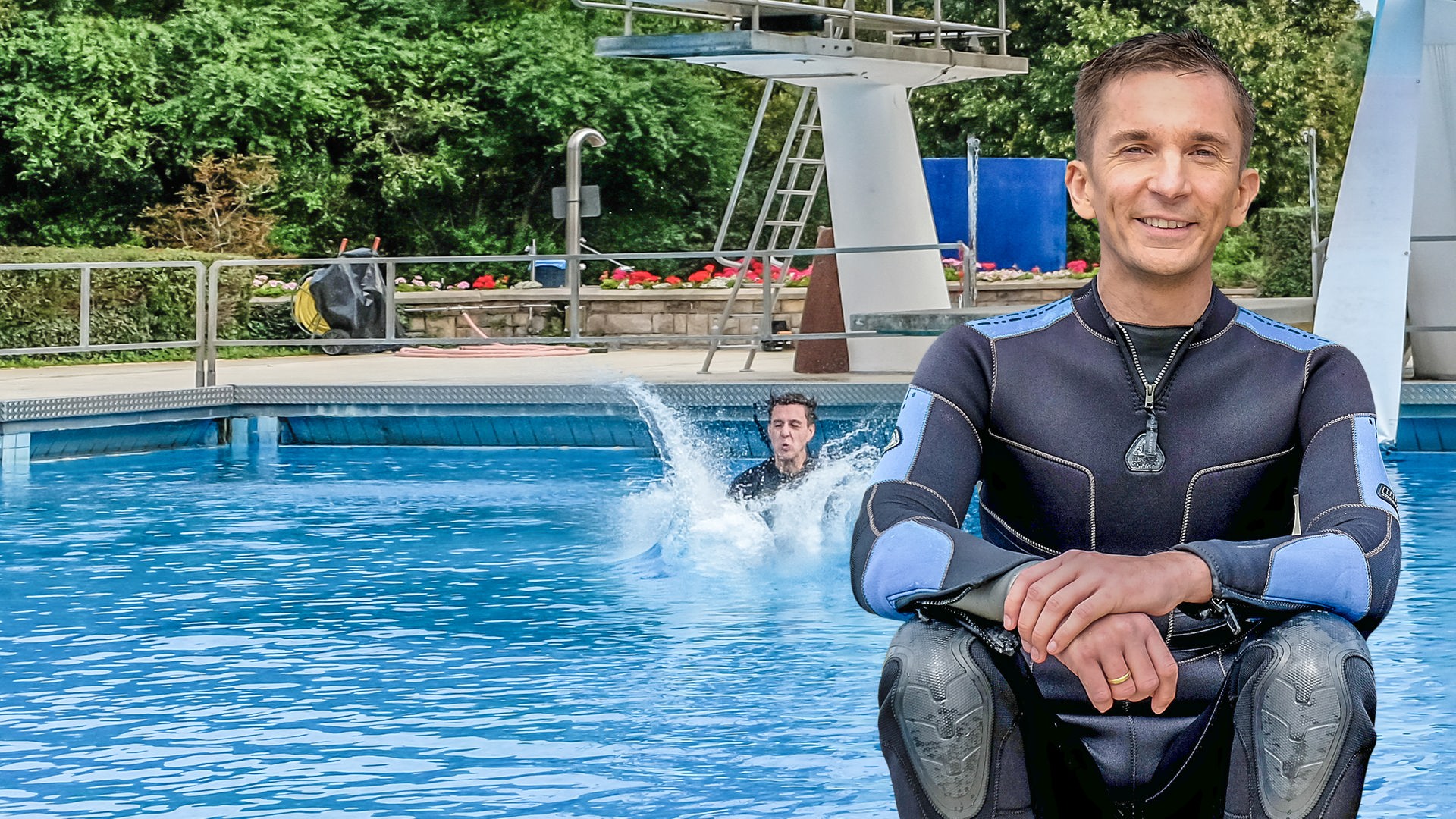 Moderator Eric Mayer sitzt im schwarzen Neoprenanzug im Vordergrund. Im Hintergrund ist ein großes Schwimmbecken mit Sprungturm. Man sieht, dass jemand im Hintergrund ins Wasser eintaucht. Wasser spritzt.
