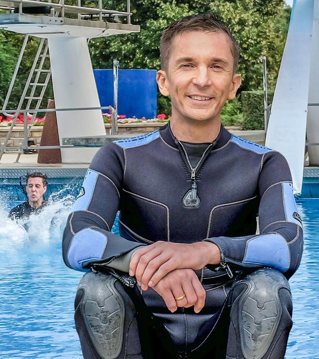 Moderator Eric Mayer sitzt im schwarzen Neoprenanzug im Vordergrund. Im Hintergrund ist ein großes Schwimmbecken mit Sprungturm. Man sieht, dass jemand im Hintergrund ins Wasser eintaucht. Wasser spritzt.