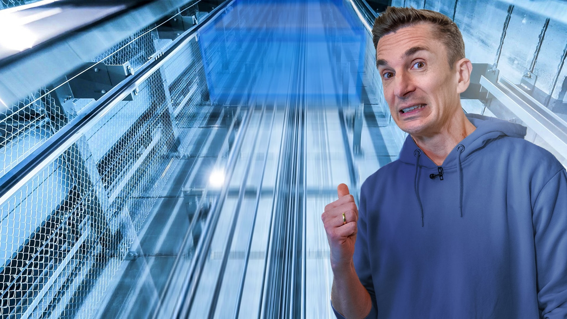 Eric im Vordergrund, schaut sorgenvoll in die Kamera. Hinter ihm befindet sich ein gläserner Aufzugschacht. 