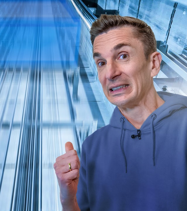 Eric im Vordergrund, schaut sorgenvoll in die Kamera. Hinter ihm befindet sich ein gläserner Aufzugschacht. 