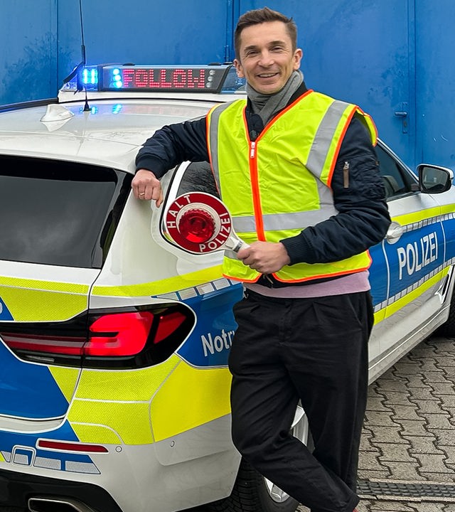 Eric steht mit Polizei-Kelle vor einem Polizeiwagen, neben ihm stehen eine Polizistin und ein Polizist.