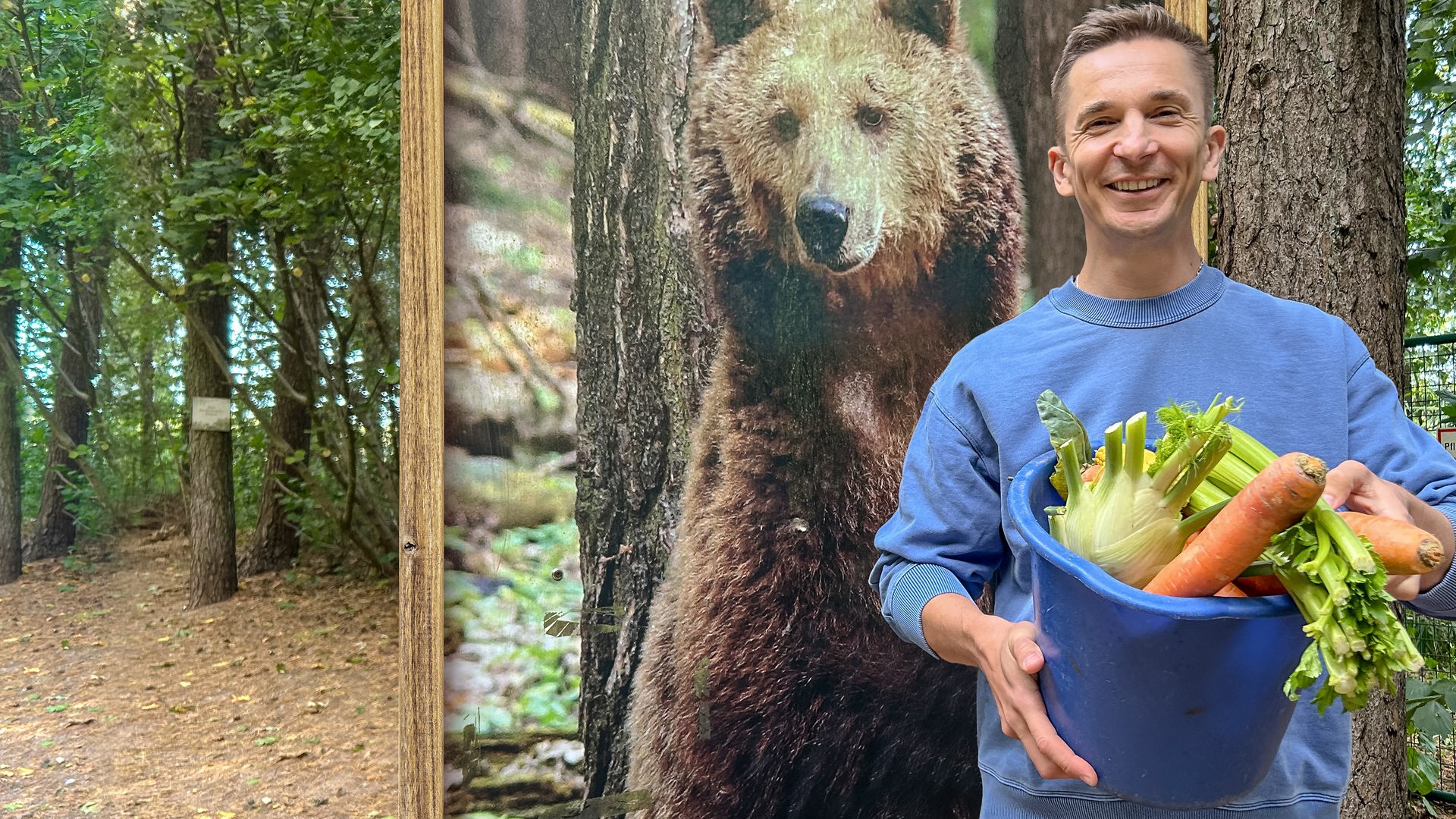 Eric steht im Wald, in der Hand hält er einen Eimer mit Gemüse, hinter ihm steht ein Aufsteller mit dem Foto eines Braunbären.