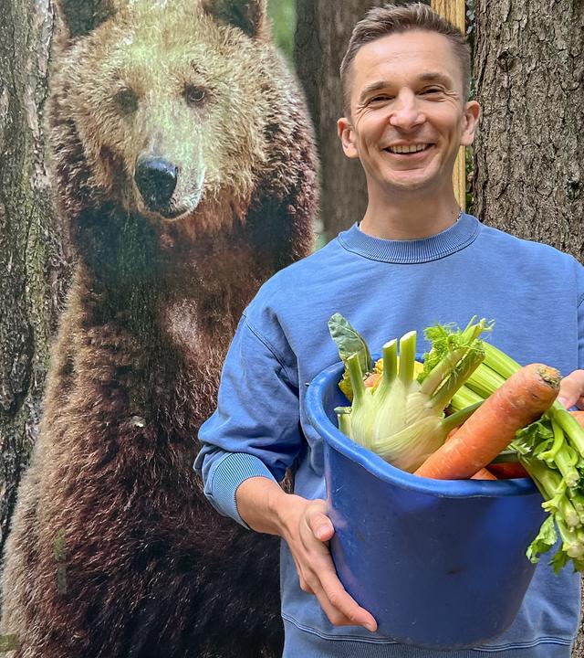 Eric steht im Wald, in der Hand hält er einen Eimer mit Gemüse, hinter ihm steht ein Aufsteller mit dem Foto eines Braunbären.