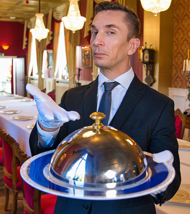 Eric als Butler in Anzug und Krawatte, in der Hand hält er eine Speiseglocke.