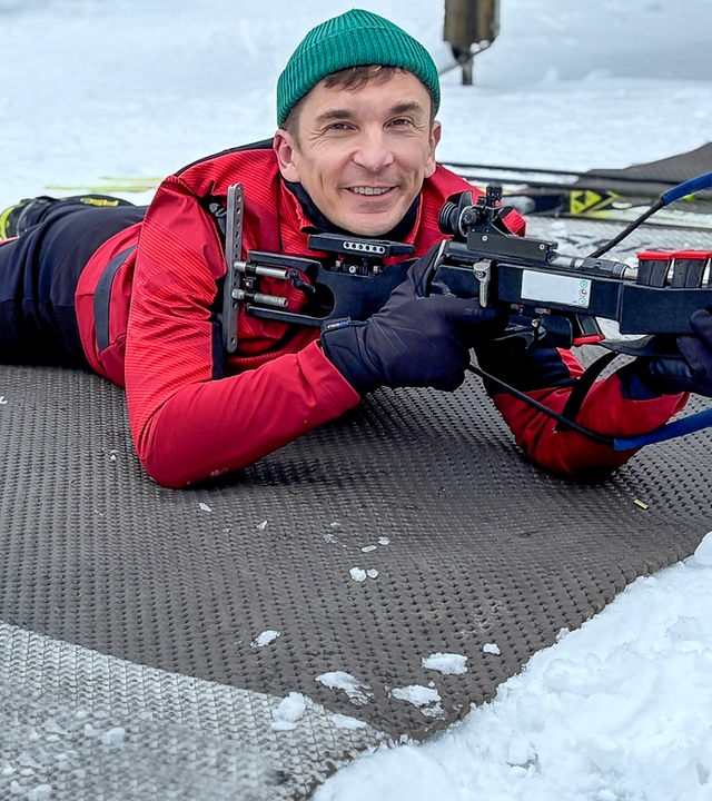 Eric Mayer liegt auf dem Boden im Schnee und bereitet sich auf einen Schuss mit dem Biathlon-Gewehr vor. Die Waffe ist auf einer Auflage stabilisiert, seine Finger liegen am Abzug. Schnee und Eis im Vordergrund machen die kalten Bedingungen sichtbar, unter denen die sportliche Aktivität stattfindet.