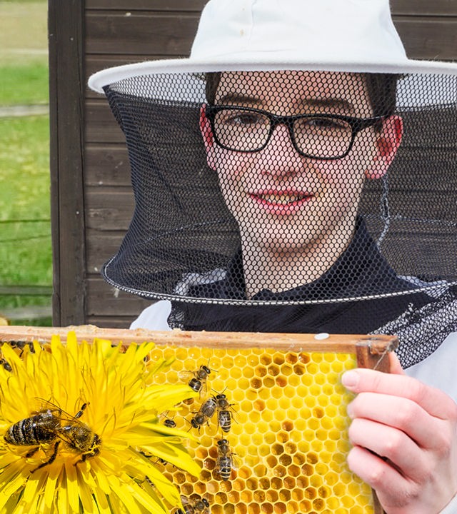 Der 13-jährige Imker Timo trägt einen weißen Schutzanzug und einen Hut mit Netz um den Kopf. In der Hand hält er eine Bienenwabe aus dem Bienenkasten mit einigen Bienen darauf.
