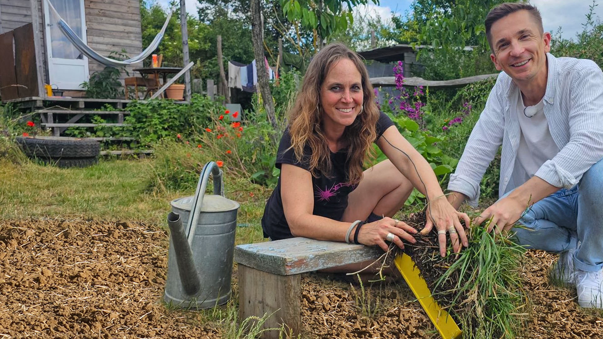 Moderator Eric Mayer kniet zusammen mit Permakultur-Designerin Johanna Häger auf dem Boden eines kleinen Gartenfeldes. Beide halten einen Büschel Gras mit Wurzeln und Erde in der Hand. Beide blicken freundlich in die Kamera.