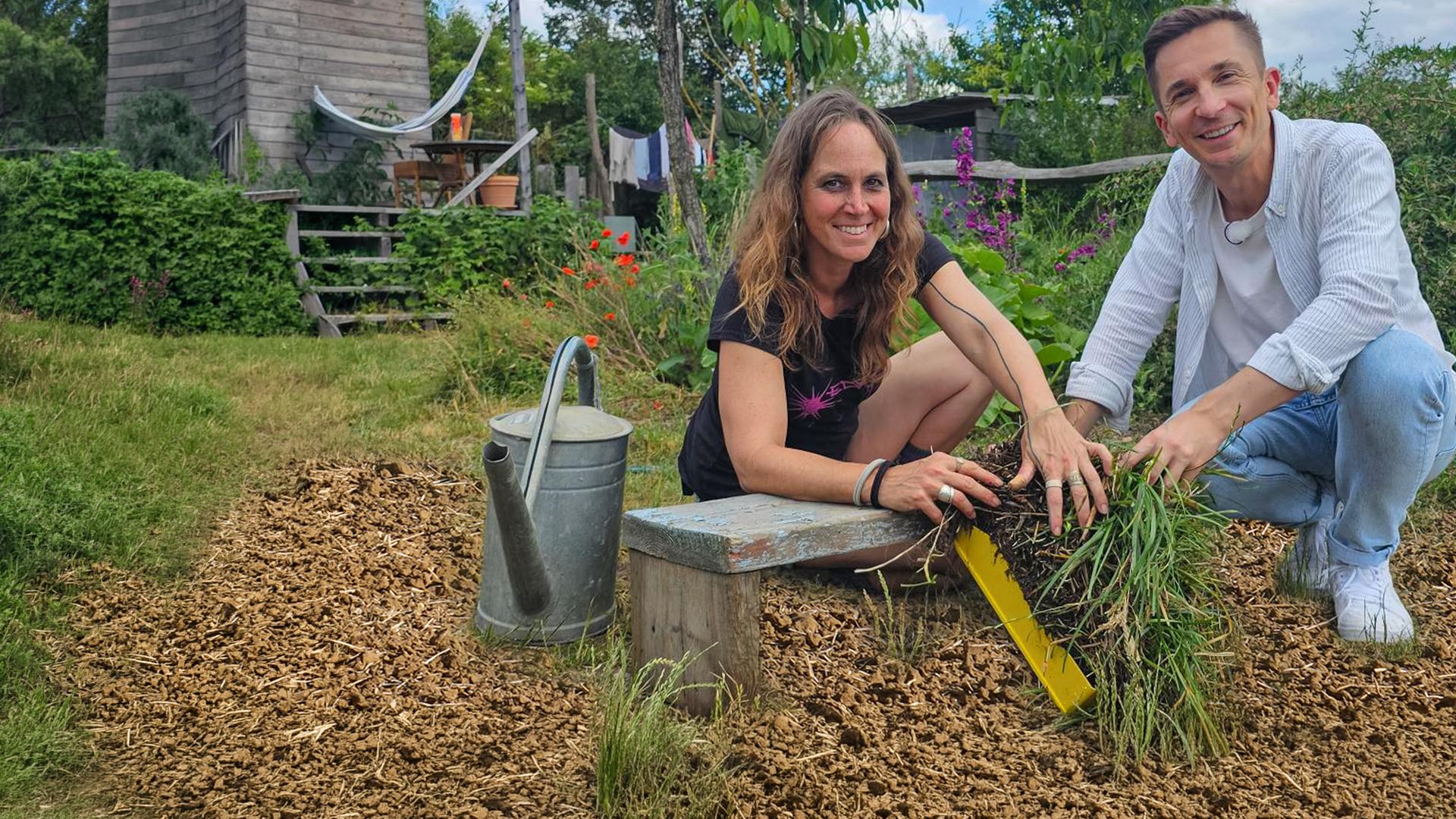 Moderator Eric Mayer kniet zusammen mit Permakultur-Designerin Johanna Häger auf dem Boden eines kleinen Gartenfeldes. Beide halten einen Büschel Gras mit Wurzeln und Erde in der Hand. Beide blicken freundlich in die Kamera.