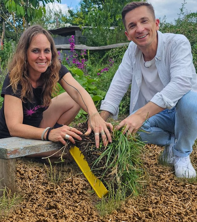 Moderator Eric Mayer kniet zusammen mit Permakultur-Designerin Johanna Häger auf dem Boden eines kleinen Gartenfeldes. Beide halten einen Büschel Gras mit Wurzeln und Erde in der Hand. Beide blicken freundlich in die Kamera.
