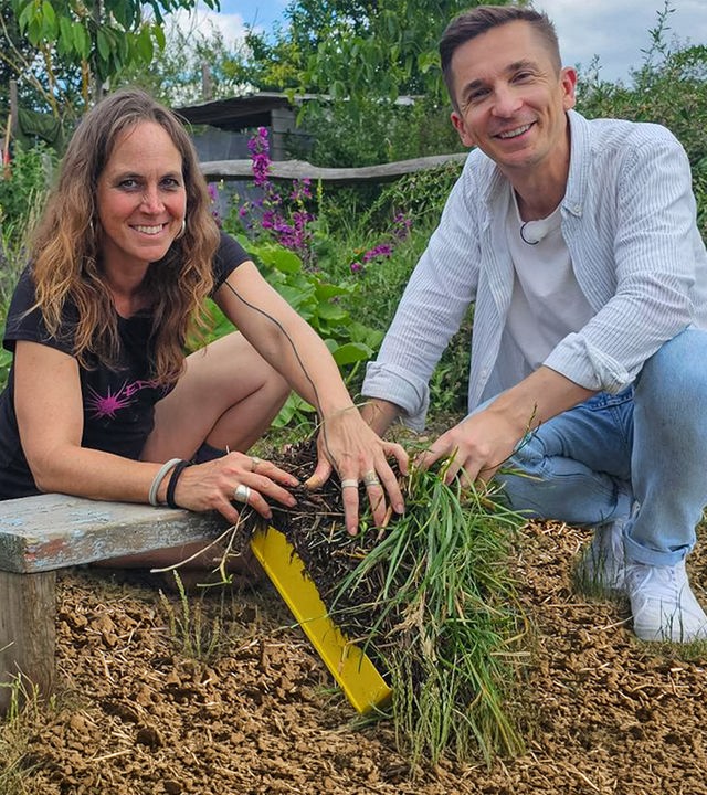 Moderator Eric Mayer kniet zusammen mit Permakultur-Designerin Johanna Häger auf dem Boden eines kleinen Gartenfeldes. Beide halten einen Büschel Gras mit Wurzeln und Erde in der Hand. Beide blicken freundlich in die Kamera.