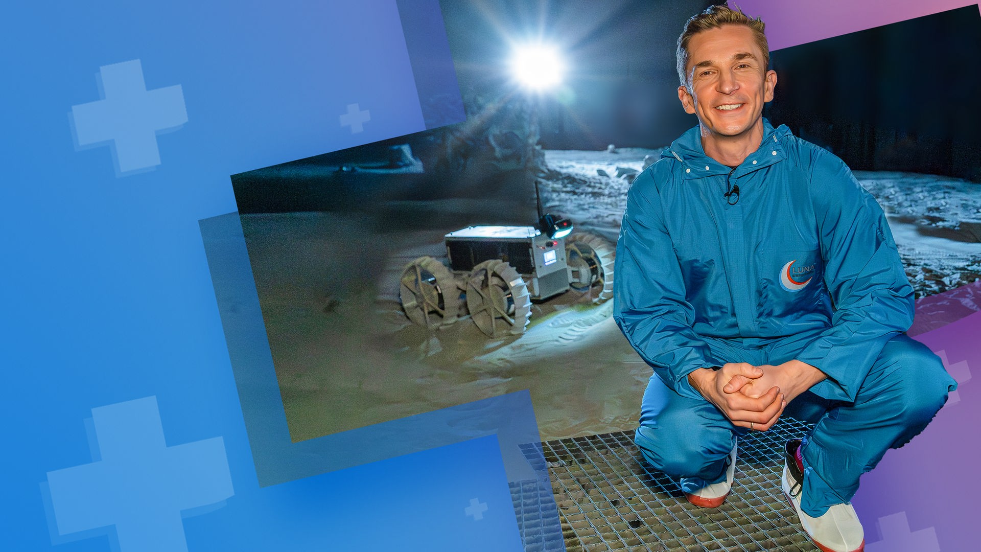 Moderator Eric Mayer hockt in der Luna Halle in Köln neben einem Mond Rover und schaut lächelnd in die Kamera. Hinter ihnm befindet sich eine Sandfläche, die von einem hell strahlenden Licht beleuchtet wird.