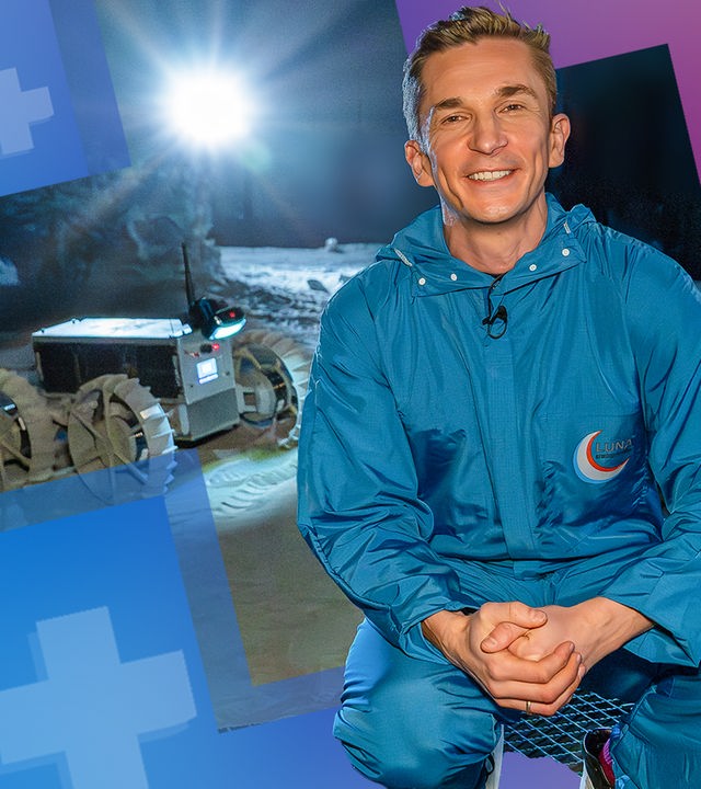 Moderator Eric Mayer hockt in der Luna Halle in Köln neben einem Mond Rover und schaut lächelnd in die Kamera. Hinter ihnm befindet sich eine Sandfläche, die von einem hell strahlenden Licht beleuchtet wird.