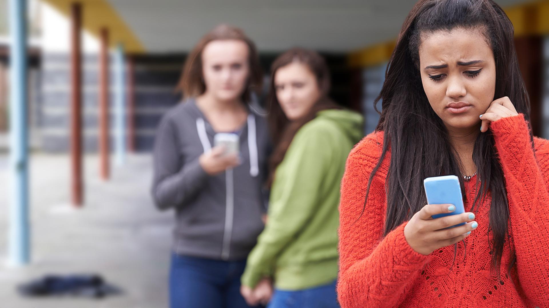 Symbolbild: Im Vordergrund steht ein Mädchen, dass traurig und frustriert auf sein Handydsplay schaut. Im Hintergrund stehen zwei Mädchen, die beide zum Mädchen im Vordergrund schauen. Eines des Mädchen hält ebenfalls ein Mobiltelefon in seiner Hand.