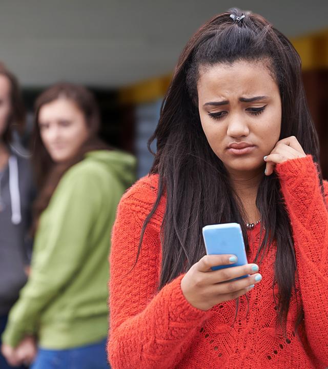 Symbolbild: Im Vordergrund steht ein Mädchen, dass traurig und frustriert auf sein Handydsplay schaut. Im Hintergrund stehen zwei Mädchen, die beide zum Mädchen im Vordergrund schauen. Eines des Mädchen hält ebenfalls ein Mobiltelefon in seiner Hand.