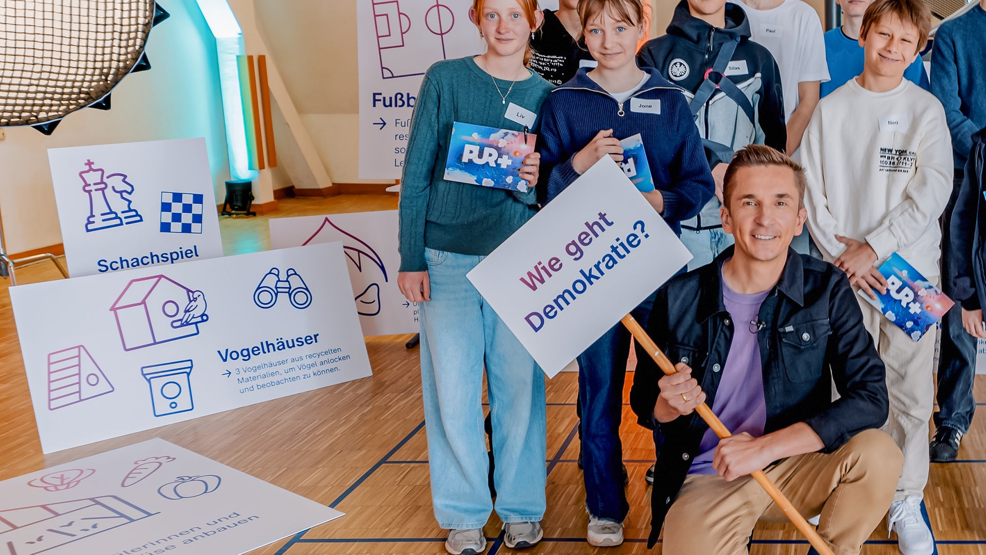 Eric steht vor einer Gruppe Schülerinnen und Schülern. Er hält ein Schild in der Hand auf dem "Wie geht Demokratie?" steht.