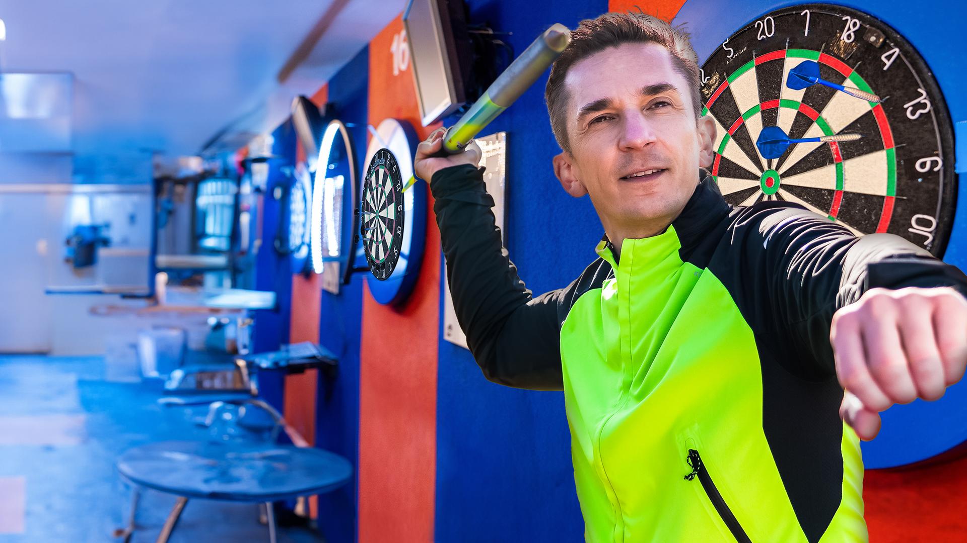 PUR+ - Eric hält einen Speer in der Hand und setzt an, ihn zu werfen. Hinter ihm befindet sich eine Dartscheibe.