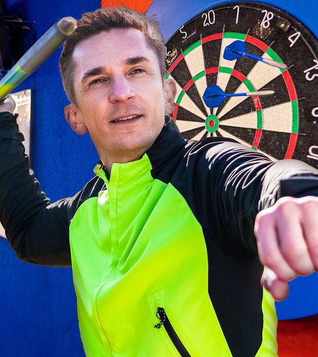 PUR+ - Eric hält einen Speer in der Hand und setzt an, ihn zu werfen. Hinter ihm befindet sich eine Dartscheibe.