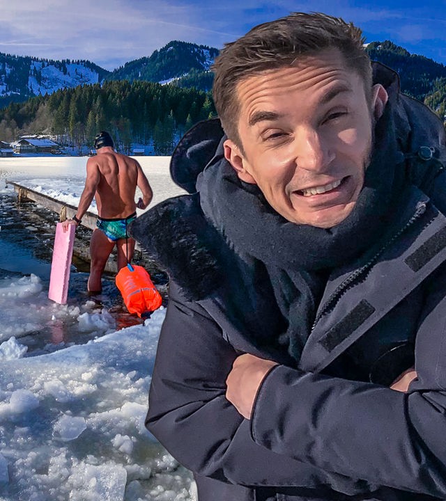 Moderator Eric Mayer steht im Vordergrund rechts. Er ist in eine dicke anthrazitfarbene Jacke eingepackt, trägt einen Schal um Hals, hält seine Arme wärmend um sich geschlungen und scheint zu frieren. Im Hintergrund geht ein Mann in Badehose ins Wasser. Rechts und links von ihm ist das Wasser zugefroren.