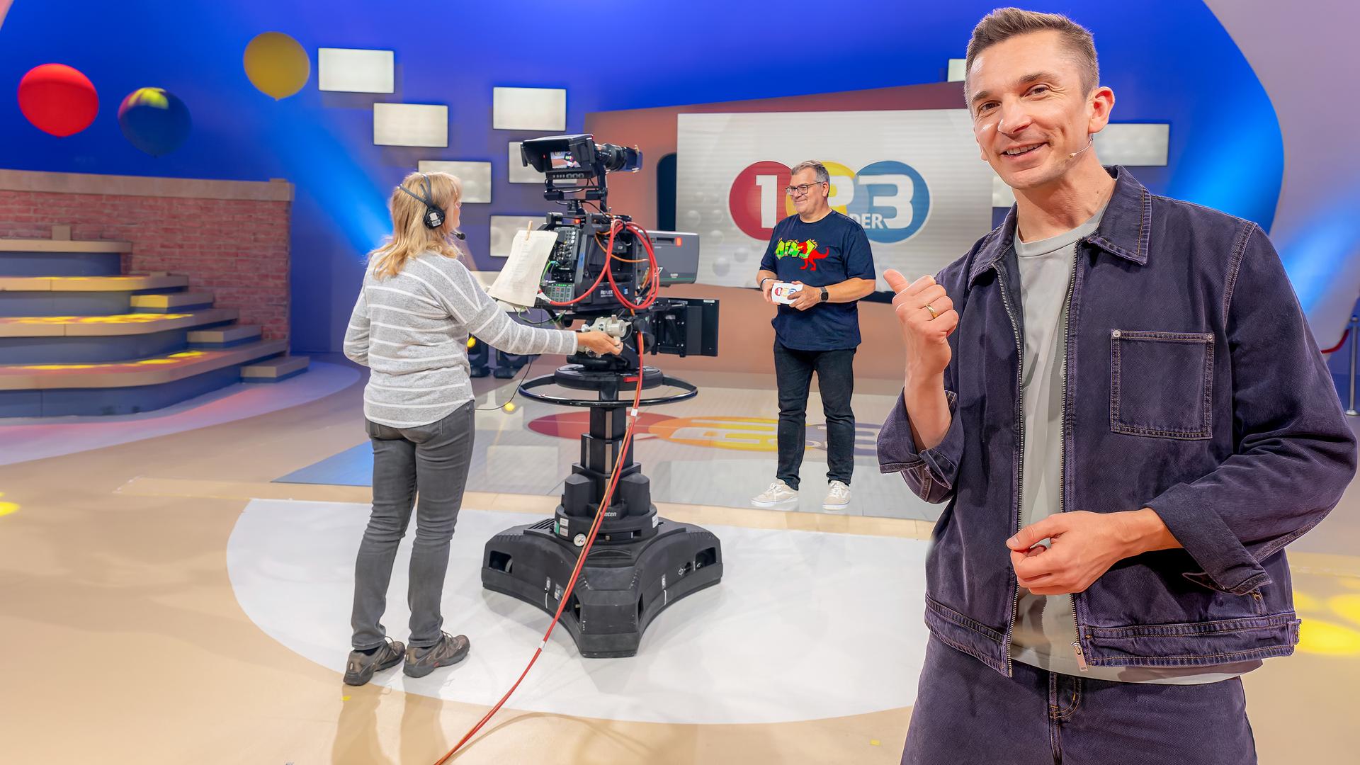 Eric schaut in die Kamera, er steht im 1, 2 oder 3 Studio, hinter ihm eine Kamerafrau an der Kamera und Moderator Elton. 