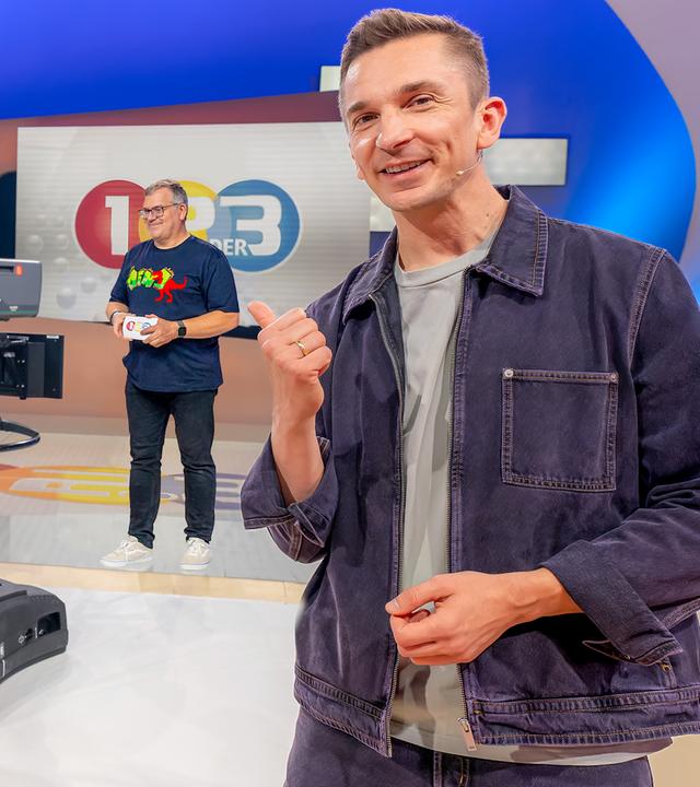 Eric schaut in die Kamera, er steht im 1, 2 oder 3 Studio, hinter ihm eine Kamerafrau an der Kamera und Moderator Elton. 