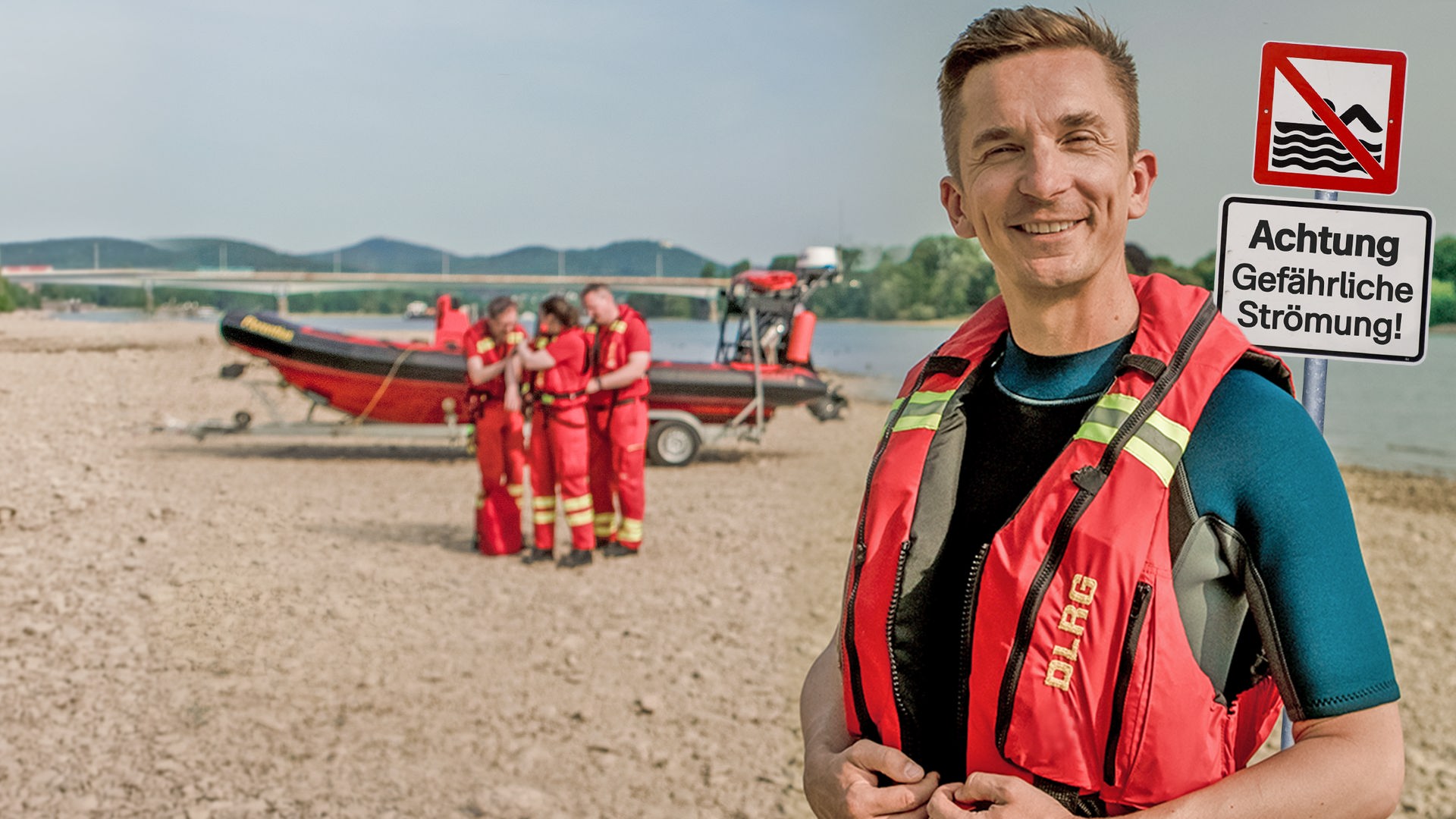 PUR+ Moderator Eric Mayer steht an einem sandigen Ufer des Flusses Rhein in einer roten Schwimmweste. Rechts neben ihm steht ein Schild mit dem Hinweis, dass das Baden hier wegen der Strömung verboten ist. Links neben ihm im Hintergrund sieht man mehrere Rettungsschwimmer an einem kleinen Rettungsboot.