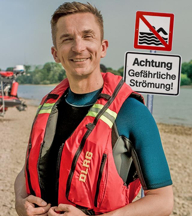 PUR+ Moderator Eric Mayer steht an einem sandigen Ufer des Flusses Rhein in einer roten Schwimmweste. Rechts neben ihm steht ein Schild mit dem Hinweis, dass das Baden hier wegen der Strömung verboten ist. Links neben ihm im Hintergrund sieht man mehrere Rettungsschwimmer an einem kleinen Rettungsboot.