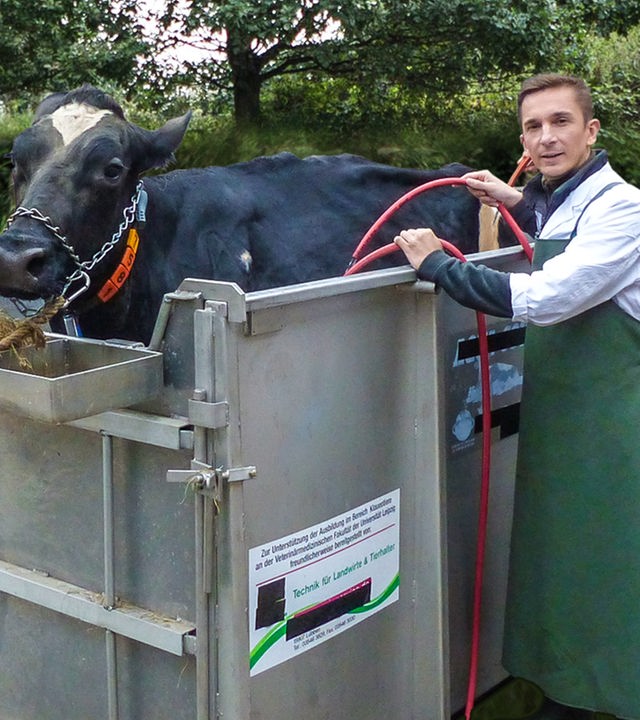 Moderator Eric Mayer steht in einem Operationssaal und schaut in die Kamera. Neben ihm steht eine Kuh, die in einem Gestell fixiert ist. Die Kuh wird gerade von einer Tierärztin, die neben Eric steht, am Magen operiert. Eric Mayer und die Tierärztin tragen grüne OP-Kleidung.
