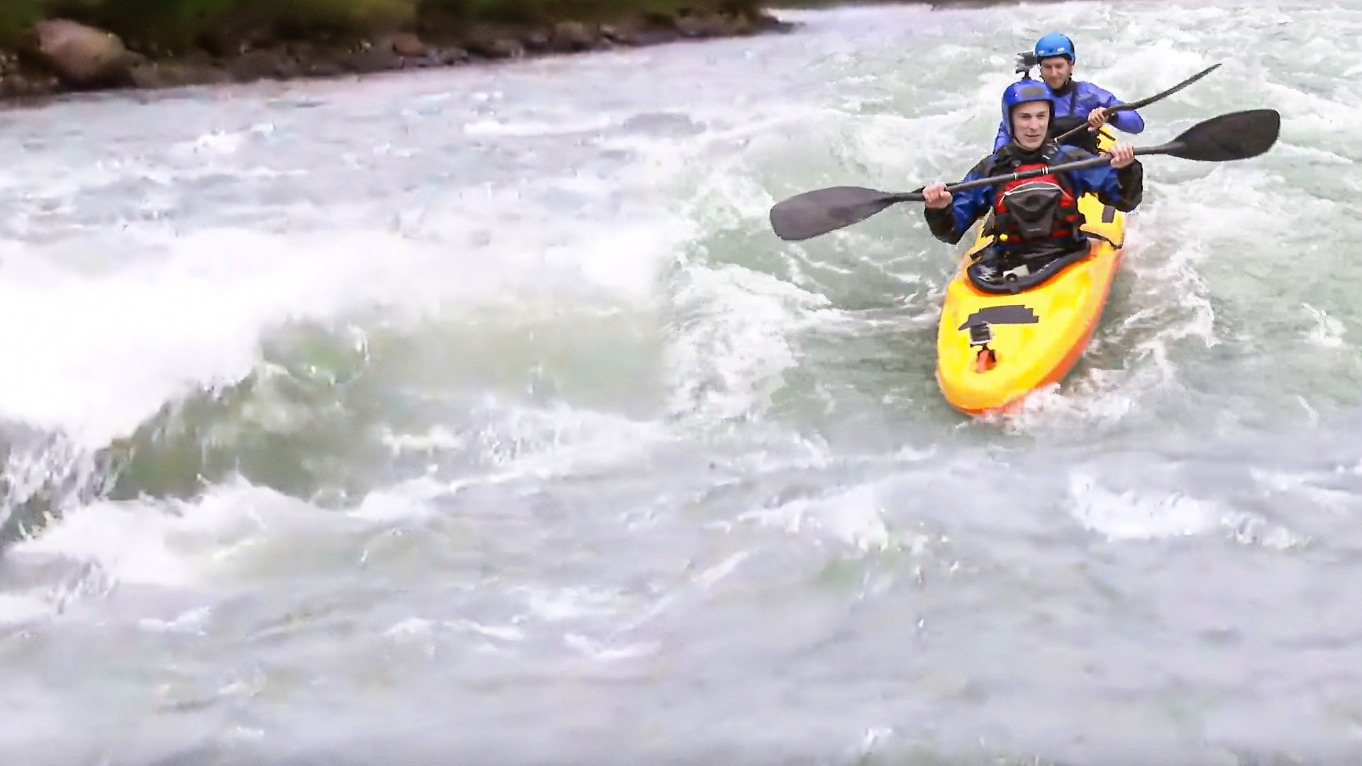 Eric Mayer sitzt in einem gelben Kajak in voller Spritzwasser-Aussrüstung und mit Helm. Hinter ihm sitzt Kajakprofi und Weltmeister Fabian Dörfler. Das Kajak fährt durch aufgewühlte Wasser. 