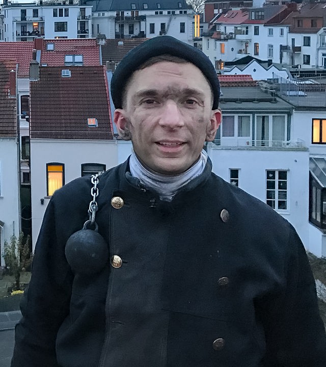 PUR+ Modertaor Eric Mayer steht in Schornsteinfeger-Arbeitskleidung und mit rußigem Gesicht in erhöhter Position und schaut in die Kamera. Im Hintergrund sind Häuser zu sehen, bei denen in manchen Fenstern der Schein einer Lampe zu sehen ist.