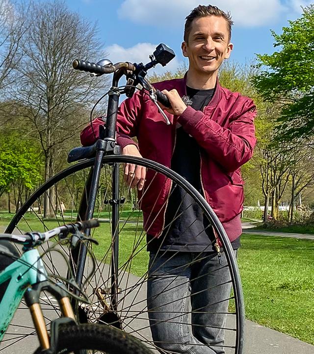  Dirtjump / Slopestyle und Freeride-Mountainbiker Lukas Knopf links, sitzend auf einem Mountainbike mit Helm und Handschuhen. Rechts daneben steht Moderator Eric Mayer angelehnt an ein altes Hochrad.