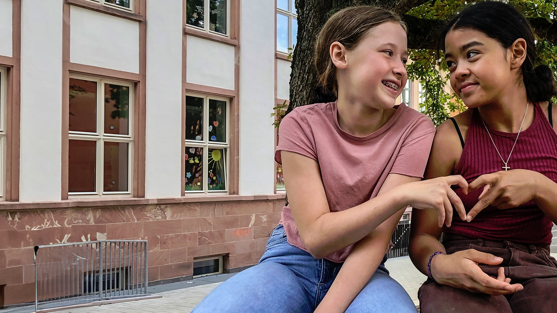 Zwei lachende Mädchen sitzen vor einem Gebäude eng beieinander und schauen sich strahlend an. Mit den Händen formen sie ein gemeinsames Herz.