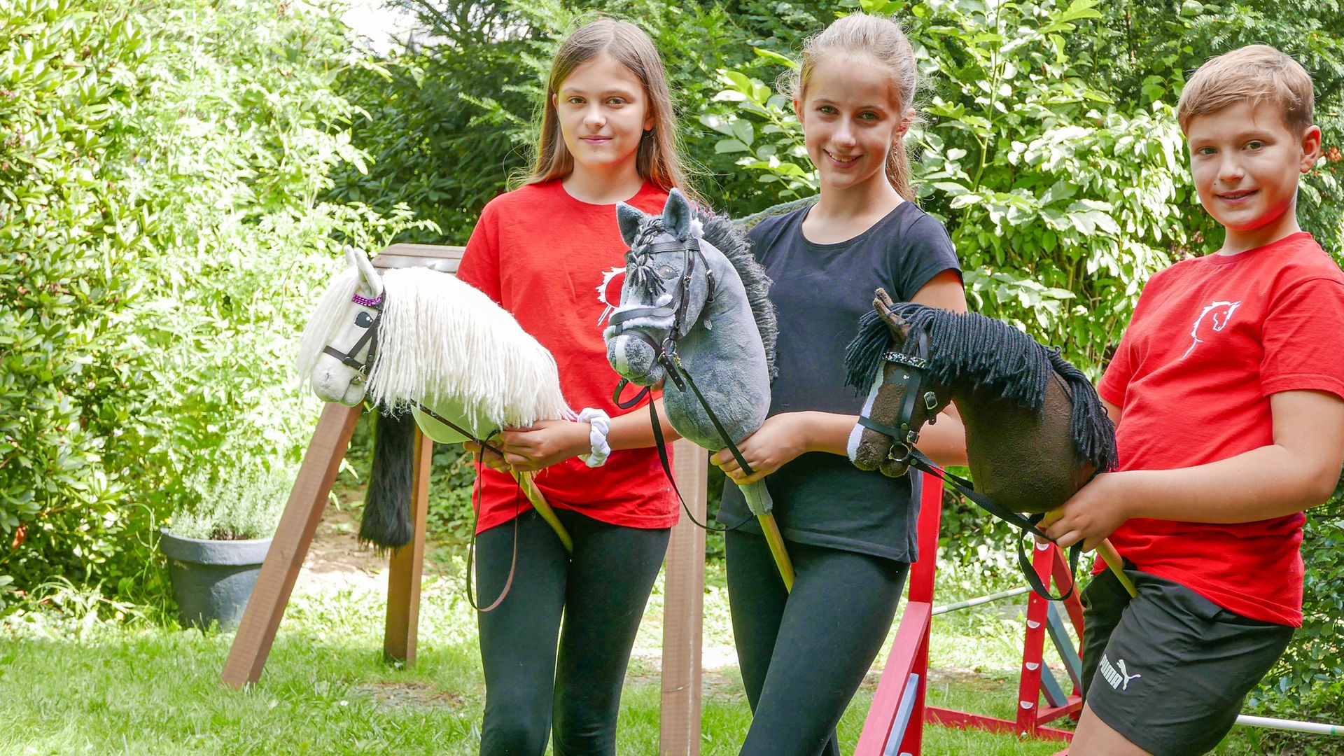 Zwei Mädchen und ein Junge stehen lächelnd nebeneinander. Ihr Hobby heißt Hobbyhorsing. Sie halten ein Steckenpferd in der Hand, dessen Stecken sie zwischen die Beine geklemmt haben. Im Hintergrund sind Teile eines aufgebauten Parcours' zu sehen.