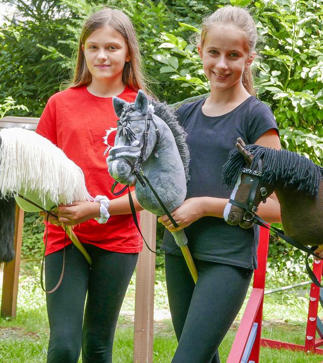 Zwei Mädchen und ein Junge stehen lächelnd nebeneinander. Ihr Hobby heißt Hobbyhorsing. Sie halten ein Steckenpferd in der Hand, dessen Stecken sie zwischen die Beine geklemmt haben. Im Hintergrund sind Teile eines aufgebauten Parcours' zu sehen.