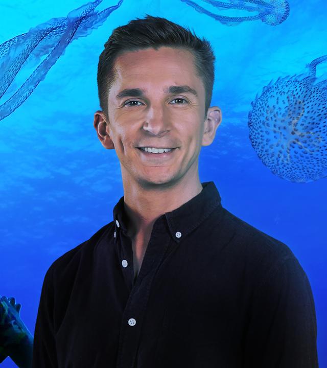 PUR+ Moderator Eric Mayer steht im Berliner Zoo vor einem riesigen Aquarium mit Quallen. Er schaut in die Kamera. Es sind schemenhaft zwei weitere Besucher zu sehen, die mit Handys ins Aquarium fotografieren.