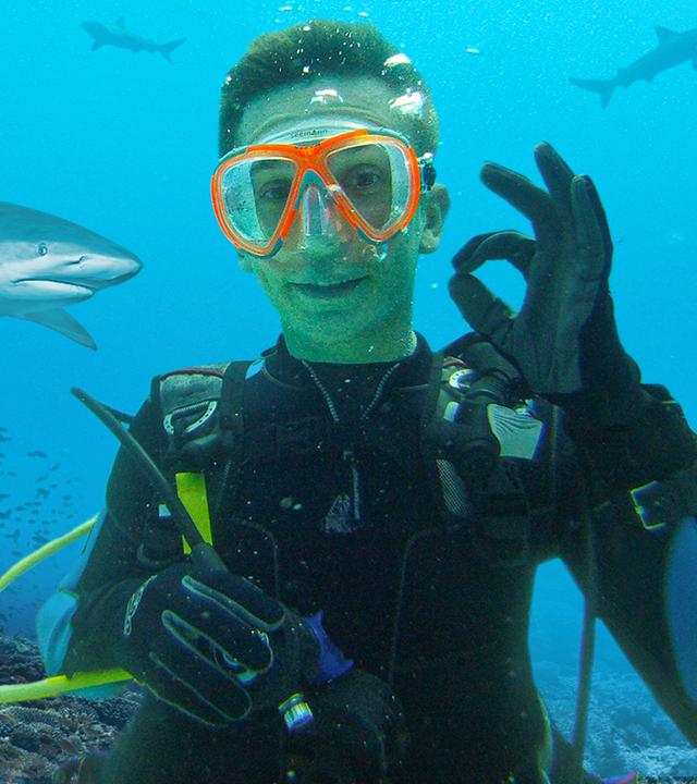PUR+ - Moderator Eric Mayer blickt unter Wasser im Taucheranzug mit Brille, Atemmaske und Atemgerät auf dem Rücken in die Kamera. Mit der linken Hand macht er das Zeichen, dass alles in Ordnung ist. Im Hintergrund sehen wir eine Unterwasser-Landschaft mit bemosten Felsen, einem Fischschwarm und einem Hai. 