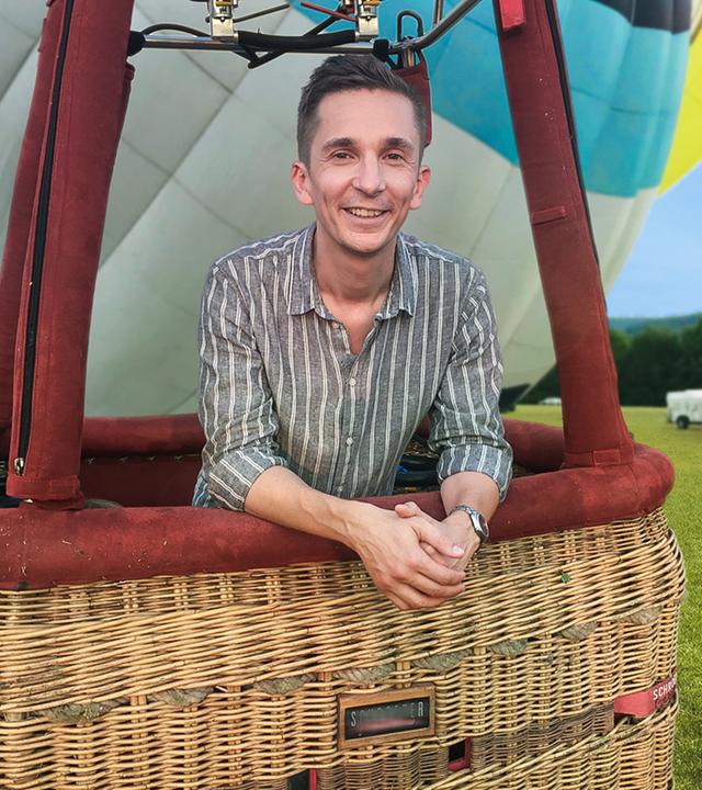 PUR+ Moderator Eric Mayer steht im Korb eines Heißluftballons, der noch auf der Erde steht. Wir sehen Eric relativ nah. Im Hintergrund liegen auf einer Wiese noch andere Ballone, die gerade aufgeblasen werden.