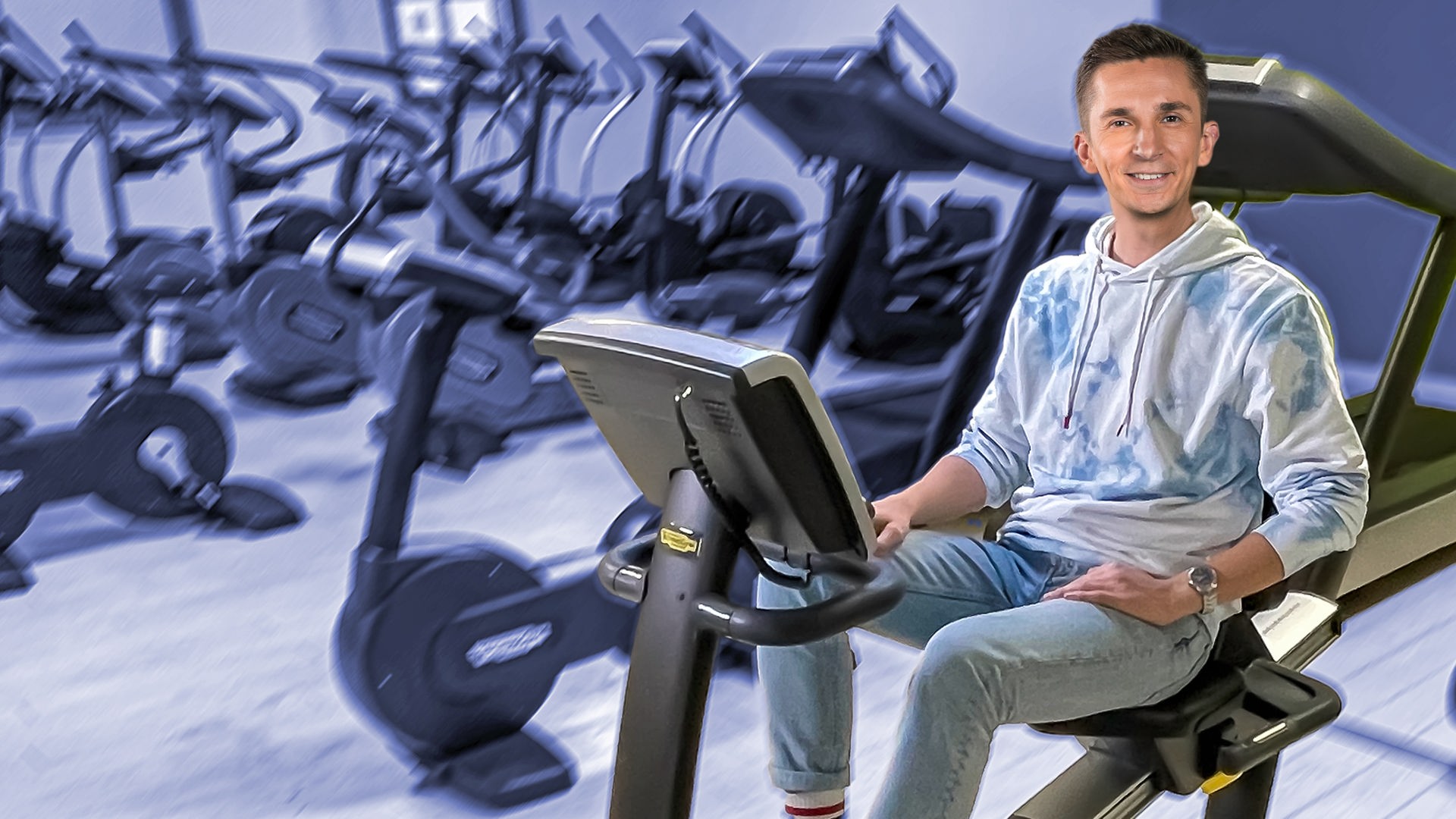 Moderator Eric Mayer sitzt auf einem Ergometer umgeben von vielen weiteren Ergometern.