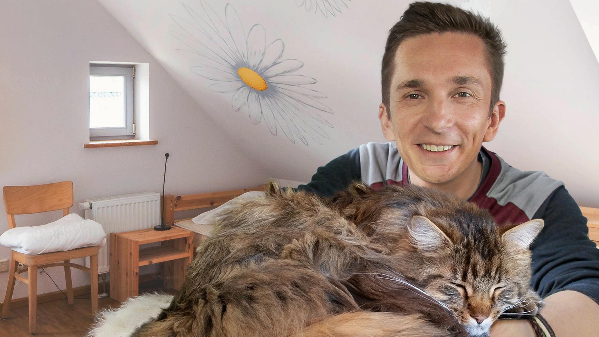 Moderator Eric Mayer lächelt in die Kamera. In seinen ineinander verschränkten Armen liegt eine Langhaar-Katze mit geschlossenen Augen und einem zufriedenen Gesichtsausdruck. Im Hintergrund ist ein Zimmer mit weißen Wänden und einem hölzernen Bett, einem Nachttisch, einem Stuhl und einem Schreibtisch zu sehen.