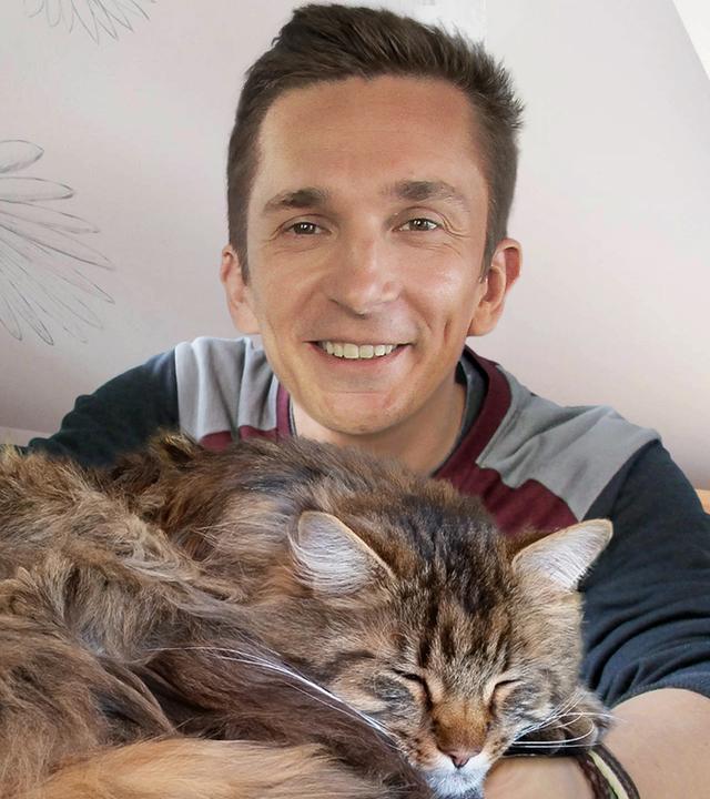 Moderator Eric Mayer lächelt in die Kamera. In seinen ineinander verschränkten Armen liegt eine Langhaar-Katze mit geschlossenen Augen und einem zufriedenen Gesichtsausdruck. Im Hintergrund ist ein Zimmer mit weißen Wänden und einem hölzernen Bett, einem Nachttisch, einem Stuhl und einem Schreibtisch zu sehen.