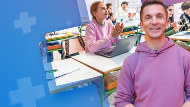 Eric Mayer steht im Vordergrund eines Klassenzimmers und lächelt in die Kamera. Hinter ihm sitzen mehrere Schülerinnen und Schüler an Schultischen, einige klatschen und wirken aufmerksam; auf einem Tisch steht ein aufgeklappter Laptop, Hefte und Stifte liegen bereit.