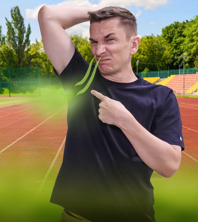 Moderator Eric Mayer hebt den linken Arm und riecht mit der Nase an seiner linken Achsel. Dabei verzieht er das Gesicht und zeigt mit dem rechten Zeigefinger auf die Achsel. Er trägt ein schwarzes T-Shirt. Im Hintergrund ist ein Sportplatz zu sehen.